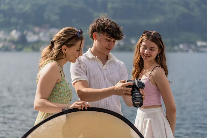 Drei Schülerinnen und Schüler der HAK Gmunden betrachten Fotos auf einer Kamera am Ufer des Traunsees.
