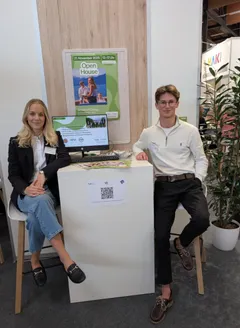 Eine Lehrerin und ein Schüler der HAK Gmunden sitzen am Messestand der Schule; im Hintergrund ein Bildschirm mit Informationen zur Übungsfirma und ein Plakat zum Open House.