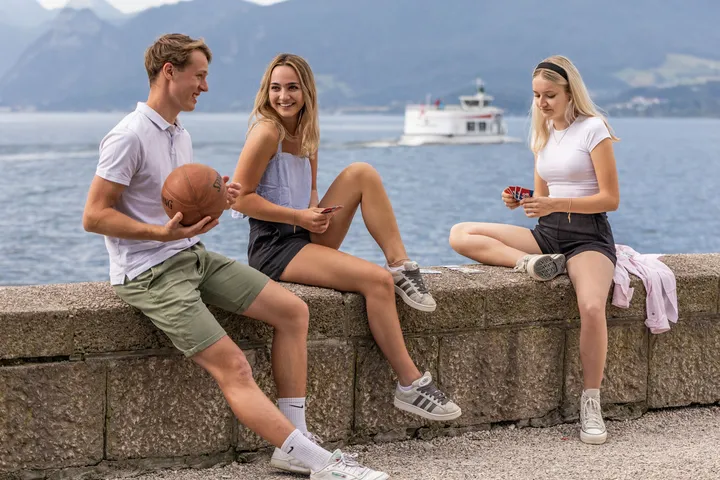 Drei Jugendliche entspannen mit Basketball und Kartenspiel auf der Seemauer am Traunsee, im Hintergrund ein Ausflugsschiff.